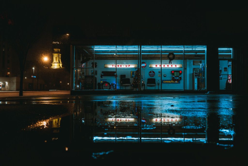 A neon-lit auto garage at night reflects on a wet pavement, creating a moody urban scene.