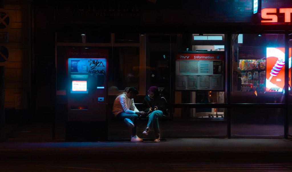 Two people sitting at a bus stop in Hamburg at night, illuminated by neon lights.