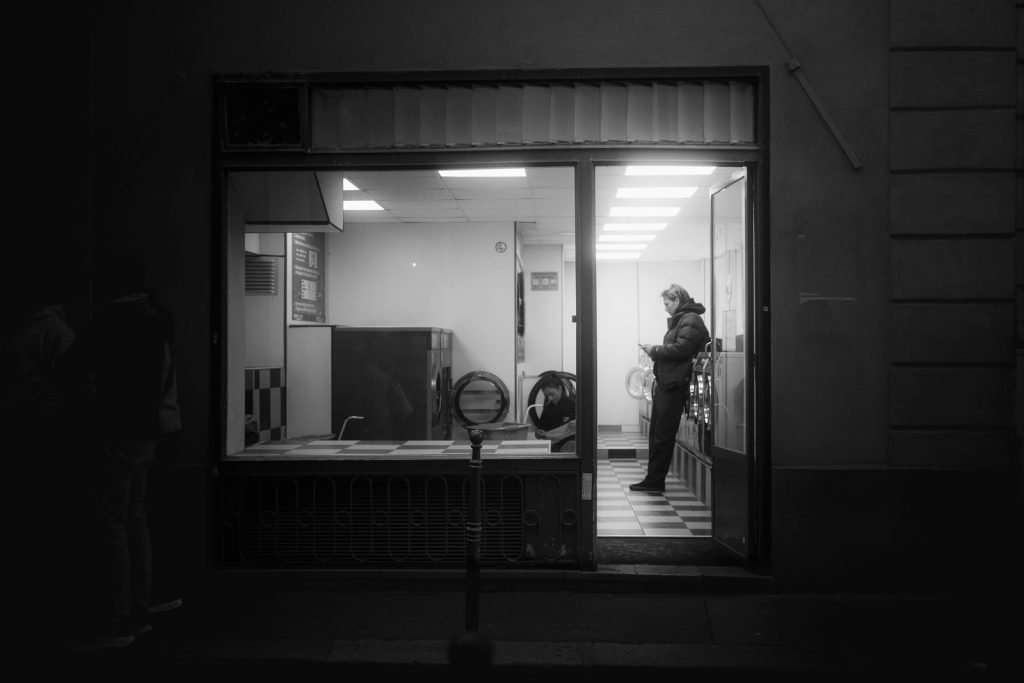 Monochrome view of a laundromat with two people inside at night, creating a moody urban atmosphere.