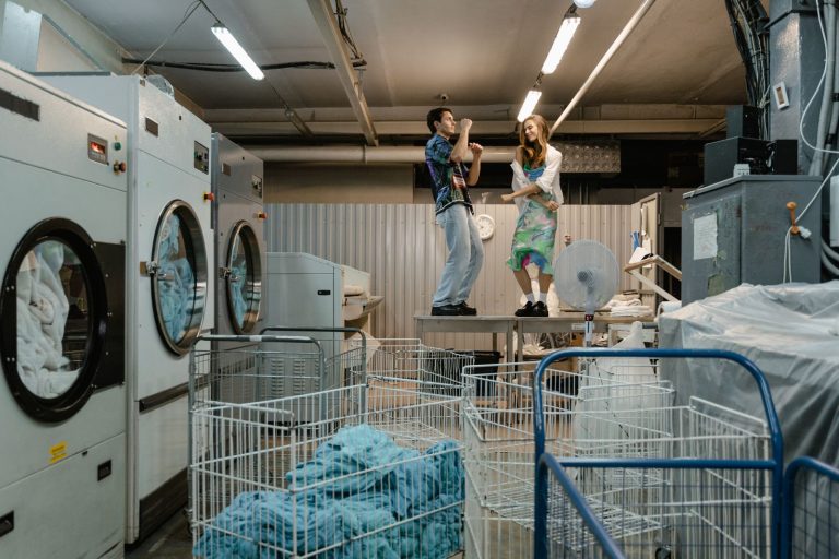 Two adults dancing energetically on a table in a modern laundromat, surrounded by industrial machines.