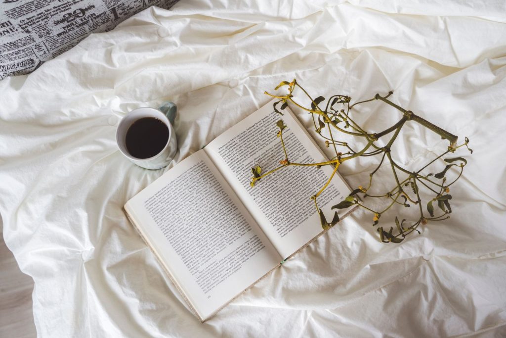 A minimalist composition of an open book, coffee cup, and dried branch on a white bed sheet.