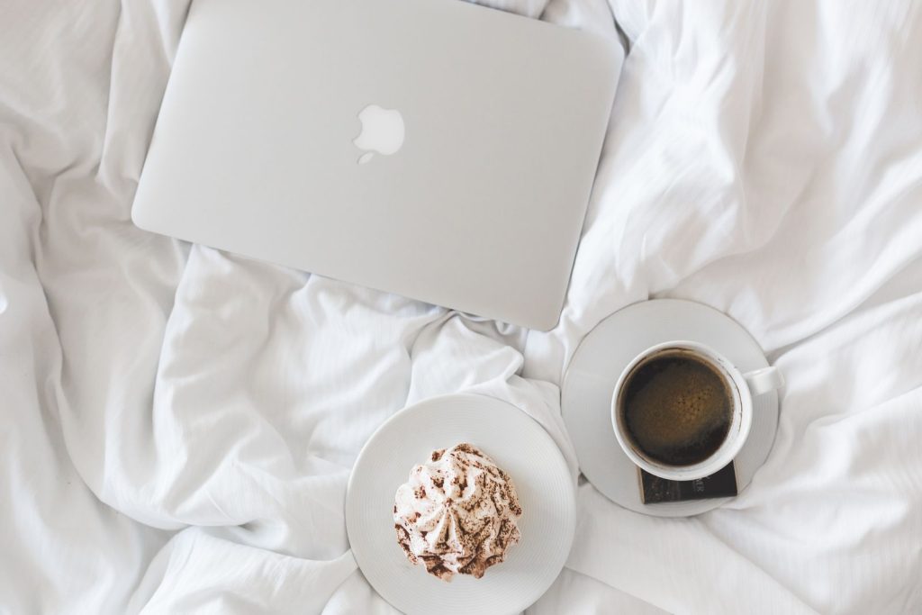 A serene morning setup with a laptop, coffee, and cake on a bed, embodying comfort and relaxation.
