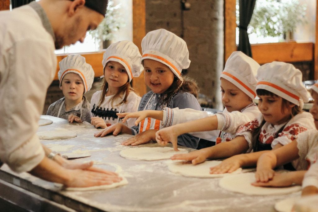 Group of children learning to bake pizza at a restaurant kitchen under a chef's guidance.