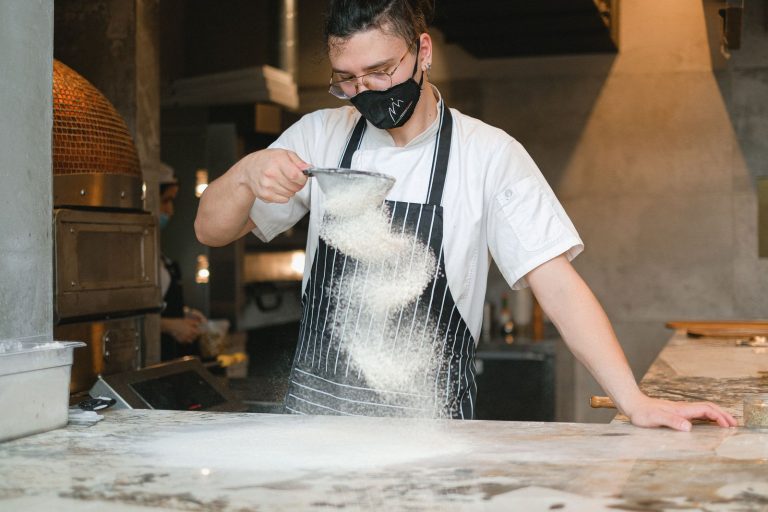 Chef in mask sifting flour in a modern kitchen, creating a fine powder effect.