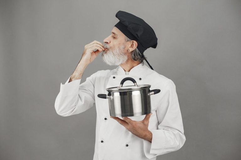Chef in uniform tasting dish with cooking pot, making hand gesture of satisfaction.
