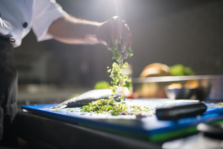 Chef's hand preparing chopped green vegetables on a cutting board in a kitchen.