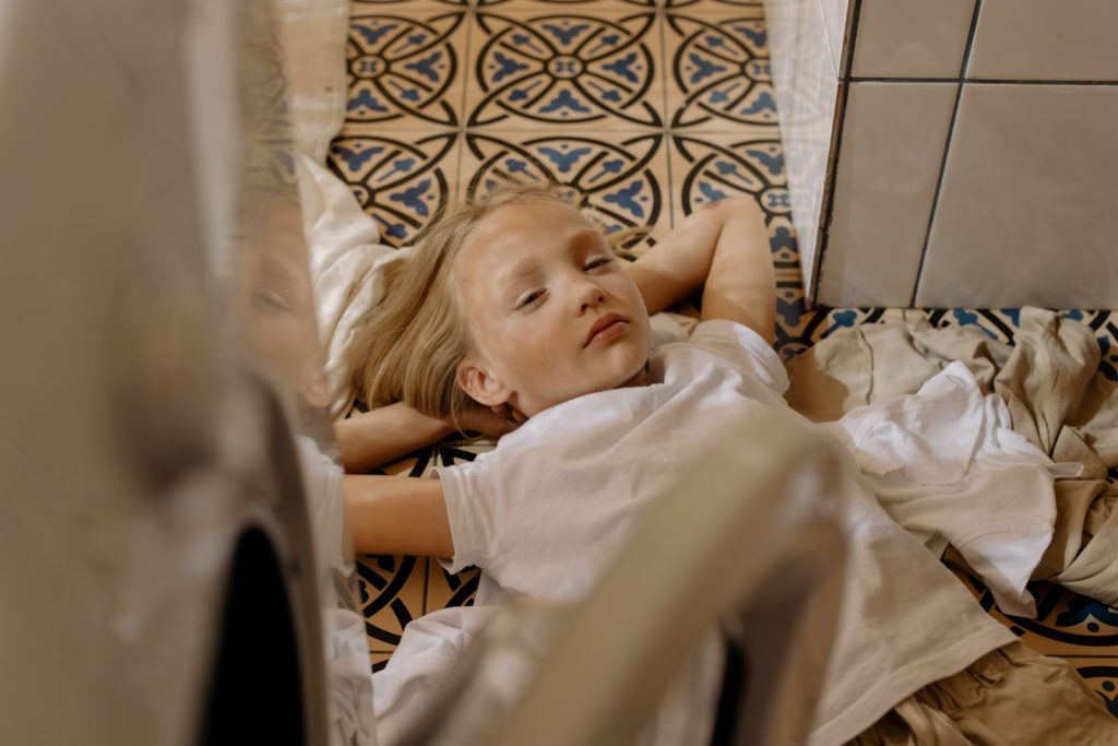 A blonde child lies comfortably on a patterned floor, surrounded by laundry indoors.