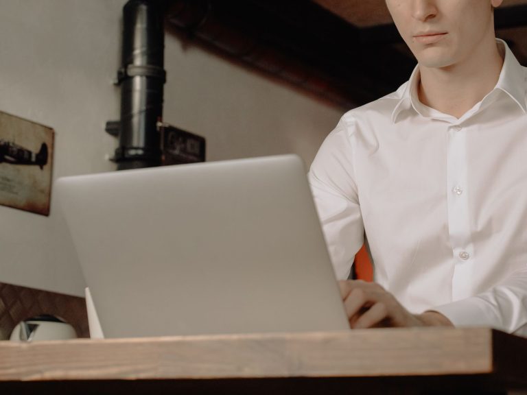Young man in a home office setting working on a laptop, dressed in a white shirt.