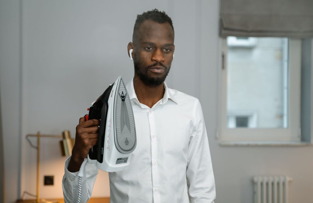 A young man in formal attire holds an iron in a home setting, wearing earbuds.
