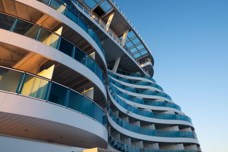 Low-angle view of a modern cruise ship with curved balconies and blue glass, under a clear sky.