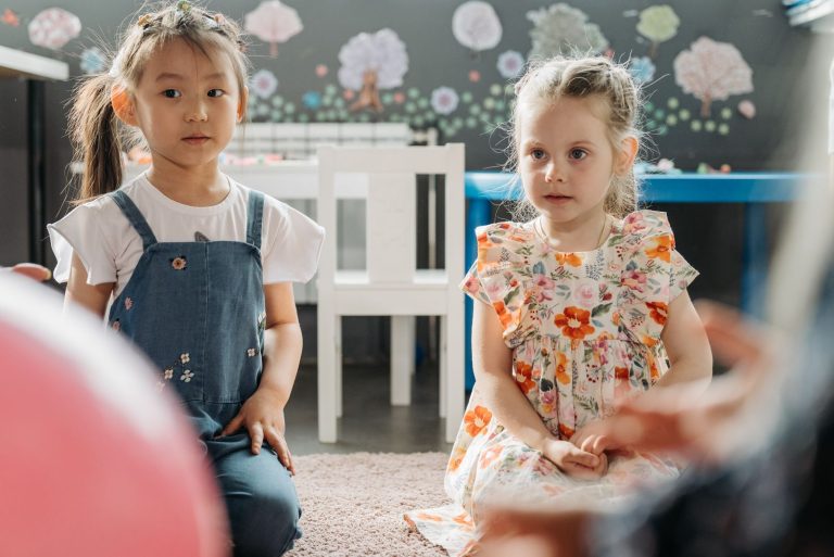 Two young girls sitting in a colorful kindergarten classroom, expressing innocence and curiosity.
