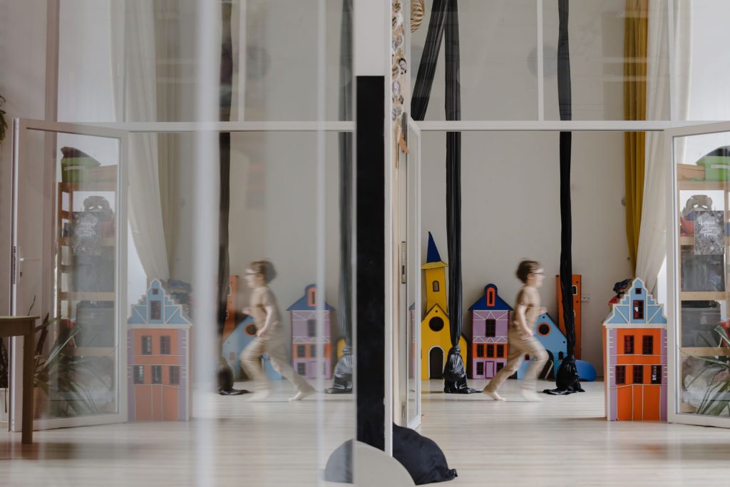 A child runs past colorful toy houses, reflecting in a glass partition, in a bright playroom.