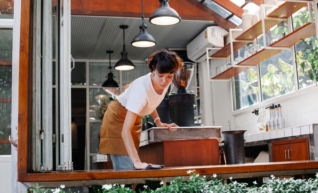 Young waitress in casual outfit and apron standing in modern cafe and washing wooden counter in daylight