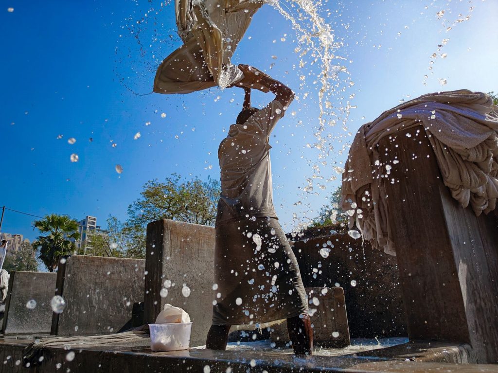 A worker vigorously washes clothes outdoors, creating a splash effect against a clear blue sky.