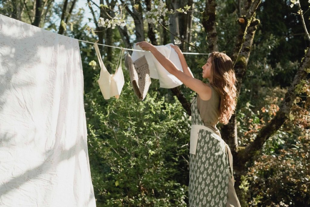 A woman hangs clothes on a line in a sunny outdoor setting, surrounded by trees.