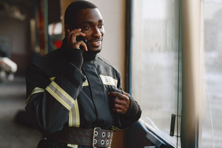 A firefighter in uniform talking on a mobile phone, depicted indoors near a window.