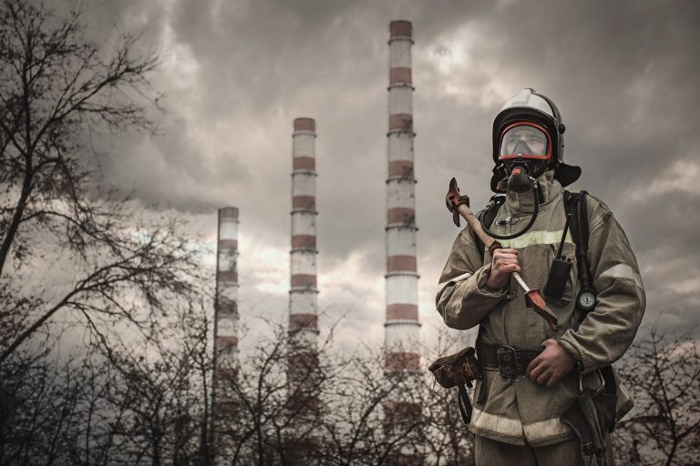 Firefighter wearing protective gear standing in front of industrial smokestacks under cloudy skies.