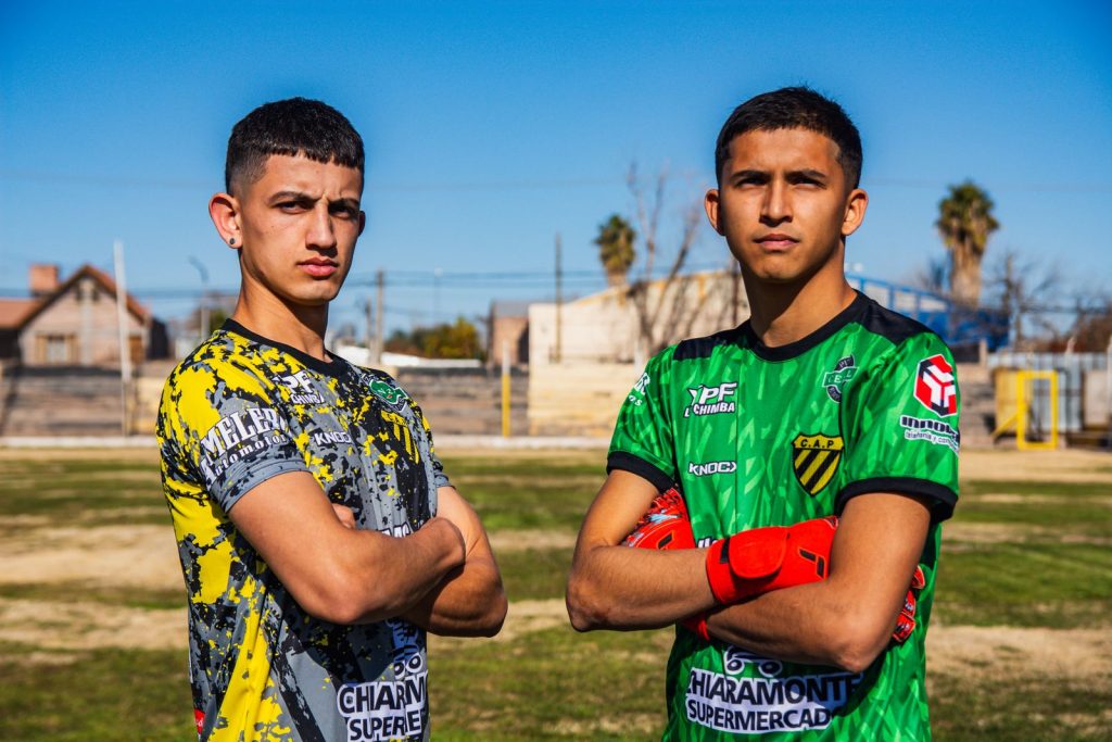 Two teenage soccer players in colorful jerseys pose confidently on an outdoor field under clear blue skies.
