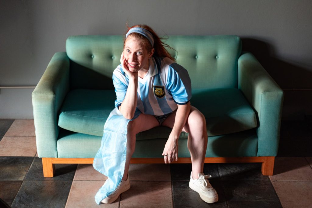A smiling woman sitting on a teal sofa wearing a blue and white Argentina football jersey indoors.