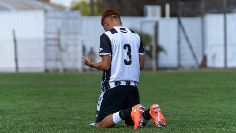 A young male soccer player kneels in prayer or reflection on the grass field.