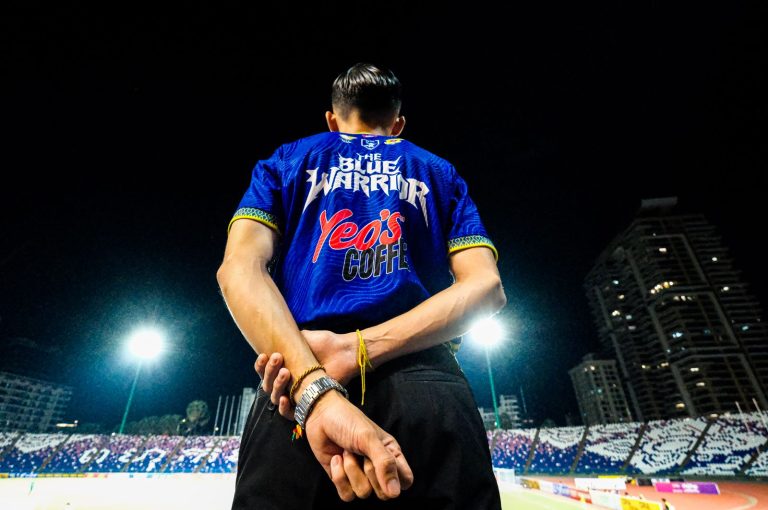 Football fan in blue jersey at night stadium stands out under floodlights, gripping anticipation.