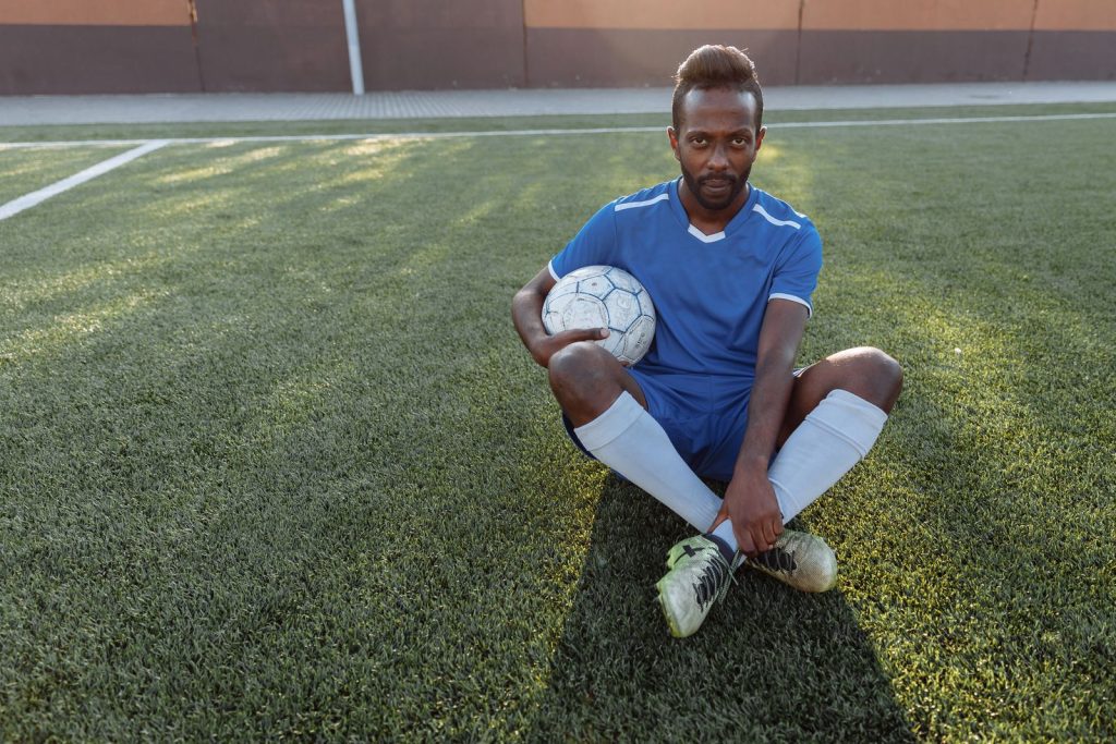 African man in blue jersey sitting on grass field holding a soccer ball.