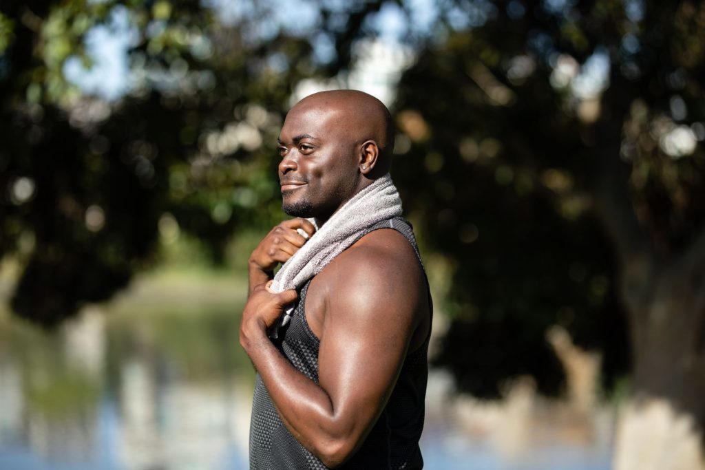 Bald man in black tank top with towel, smiling outdoors with blurred background.