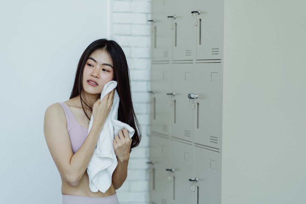Woman in a locker room drying off with a white towel, next to lockers.