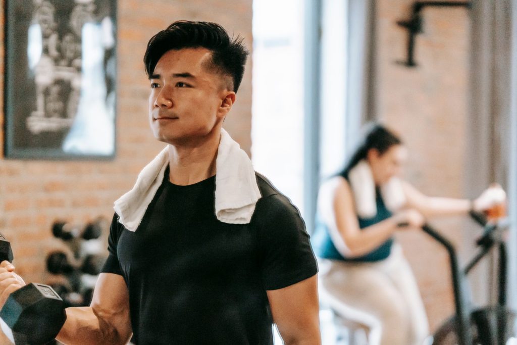 Determined Asian male athlete with towel lifting heavy dumbbell while training in spacious gym with plump woman on blurred background