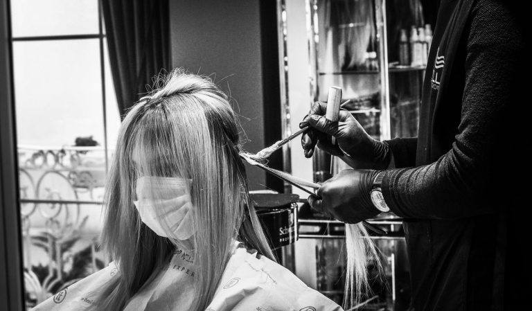 Hair stylist applying color to a woman wearing a mask in a salon, in black and white.