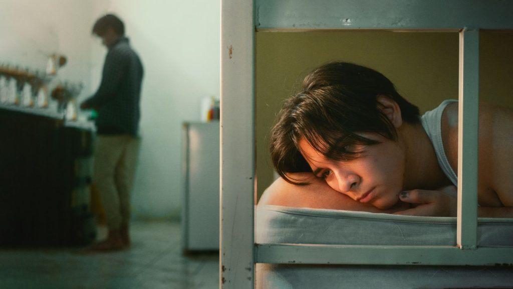A young man lying on a bed looking pensive indoors. Another person is in the background.