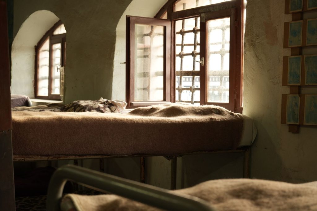 Sunlight streams through open windows onto a cozy bed in a rustic room, creating a warm atmosphere.