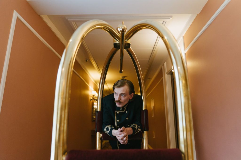 Hotel bellhop dressed in a vintage uniform leaning on a luggage trolley in a brightly lit hallway.