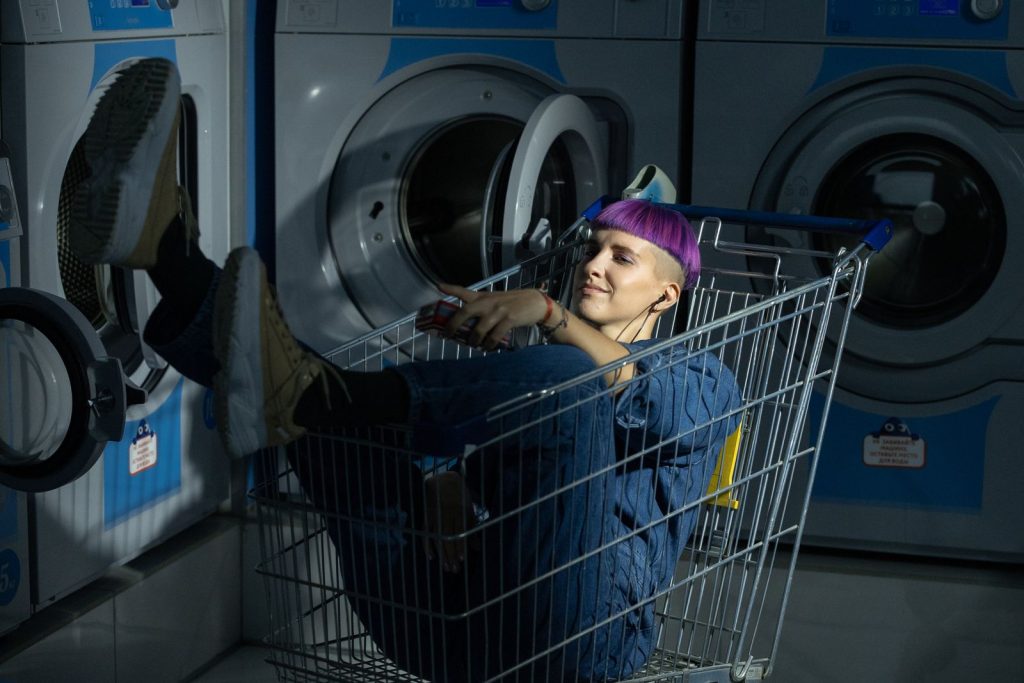 Young adult with dyed hair lounging in a shopping cart at a laundromat, embracing a quirky style.