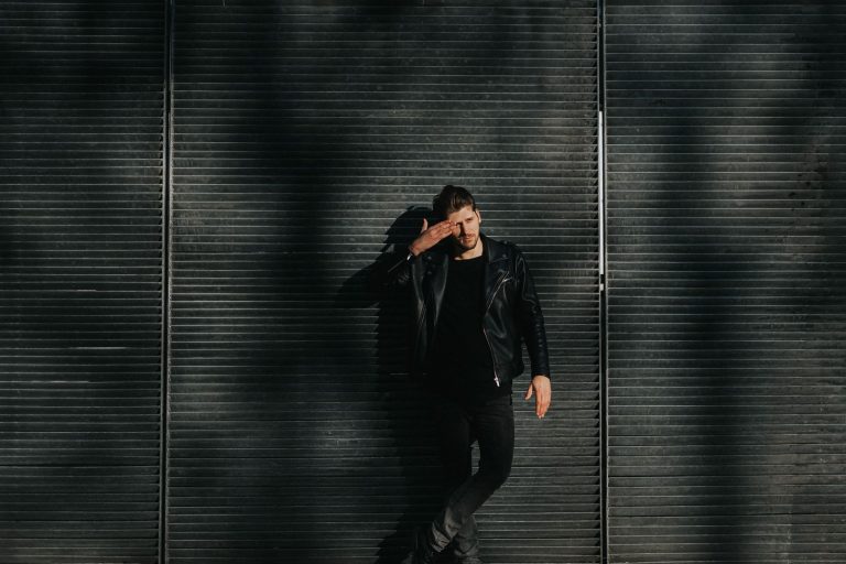 Young man poses confidently in a black leather jacket against a gridded metal urban wall.