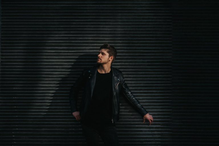Fashionable adult male in black leather jacket posing against textured wall in moody light.