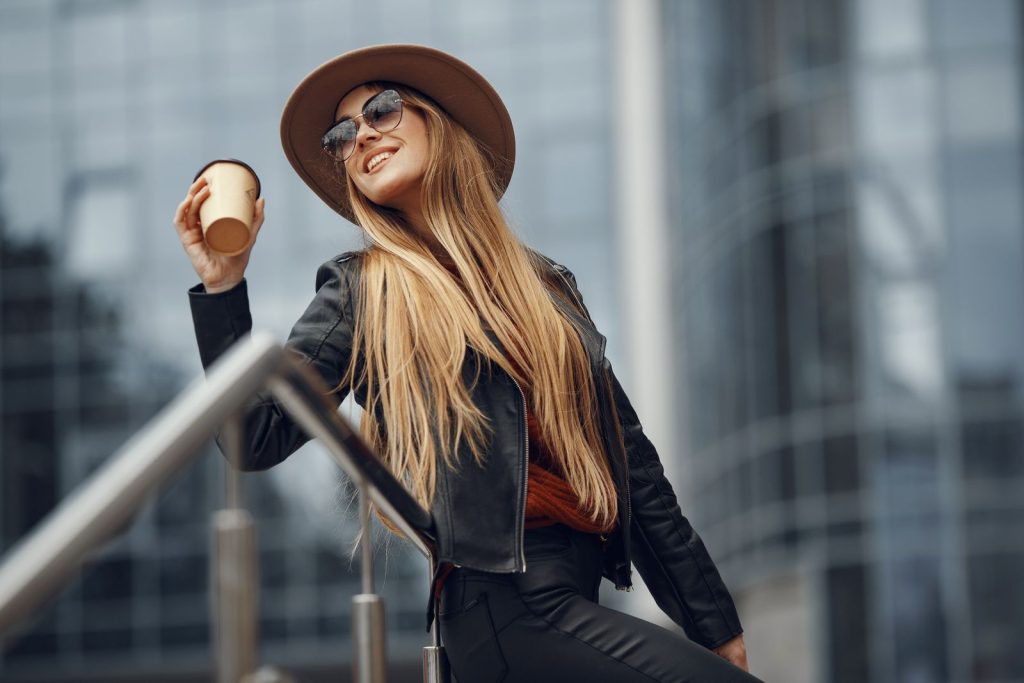 Fashionable woman in leather jacket and hat holding coffee cup in an urban environment.