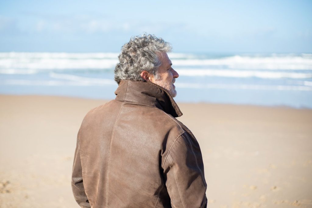 Senior man gazing at ocean on Portugal's sandy beach, wearing leather.