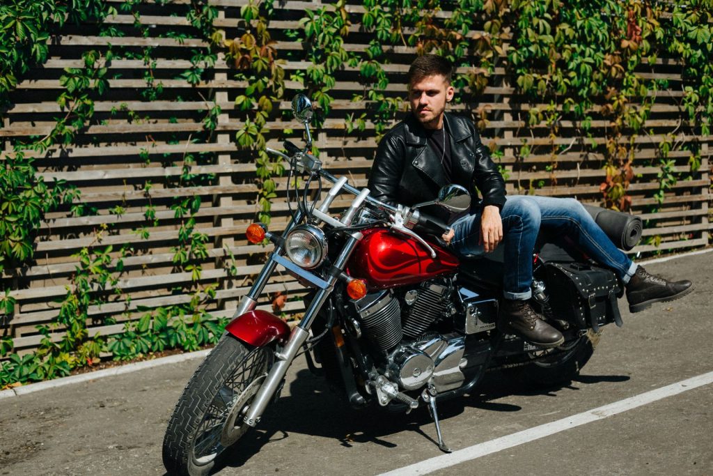 Man in leather jacket sitting on a motorcycle outdoors against a wooden fence with greenery.