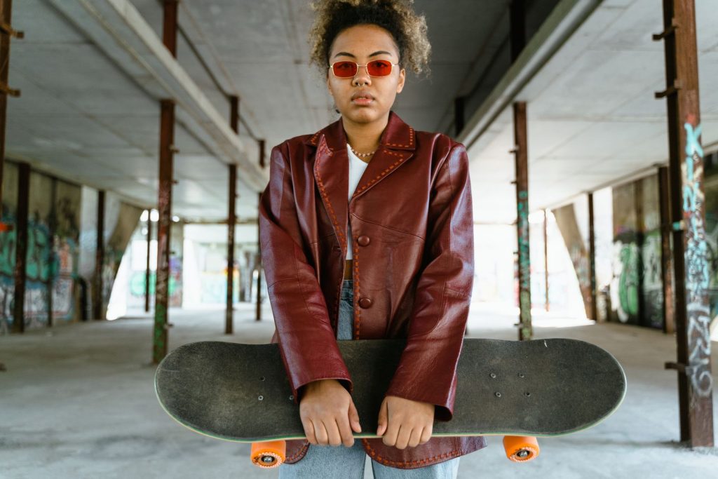 A young woman in a red leather jacket and sunglasses holds a skateboard in an urban graffiti-filled space.
