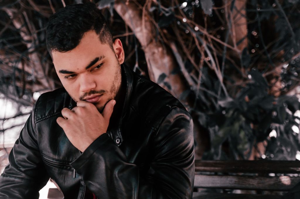 A thoughtful man in a leather jacket rests his chin on his hand outdoors with a blurred background.