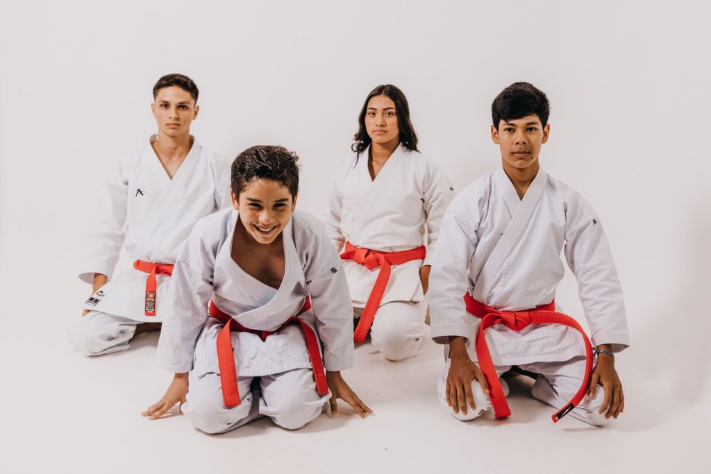 Four teenagers in martial arts uniforms showcase their skills with red belts on a white background.