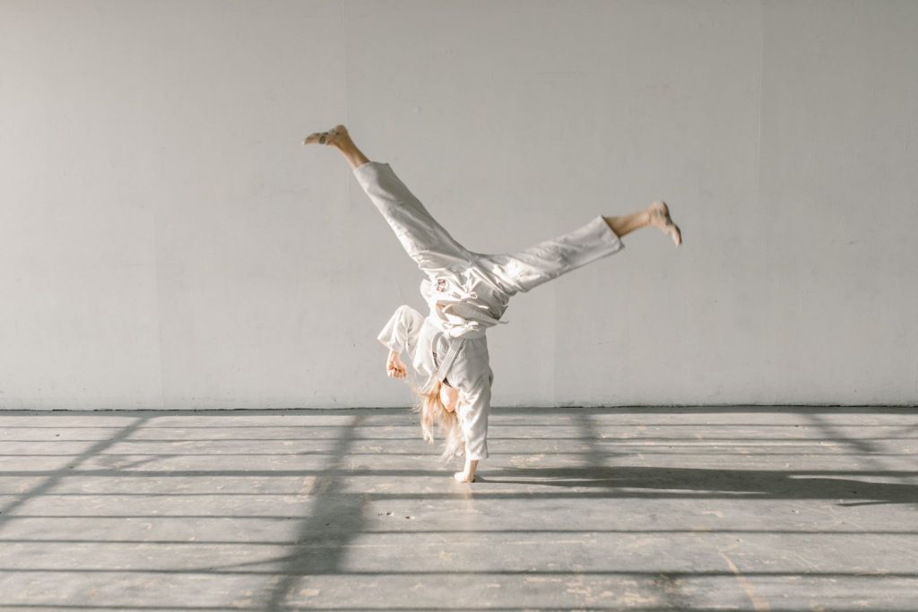 A woman in a white gi performing a handstand in a well-lit gym setting.