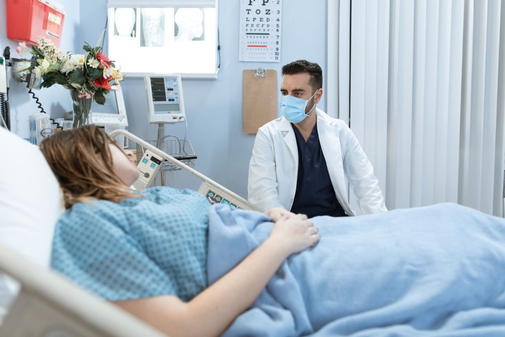 A doctor in a face mask talks to a patient in a hospital bed, providing care and consultation.