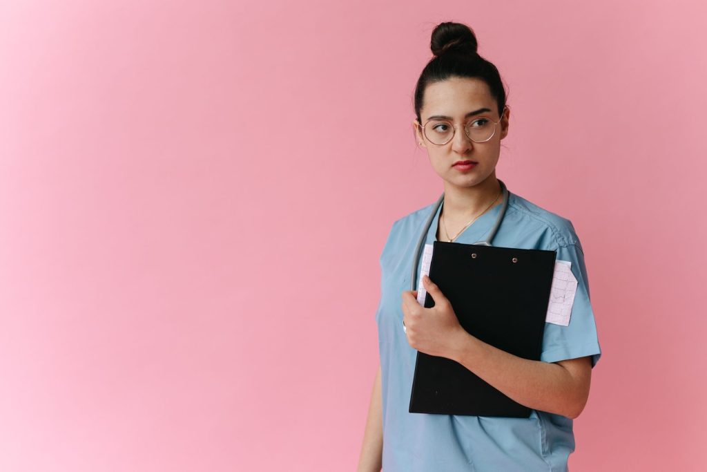 Confident female doctor in scrubs holding a clipboard against a pink background.