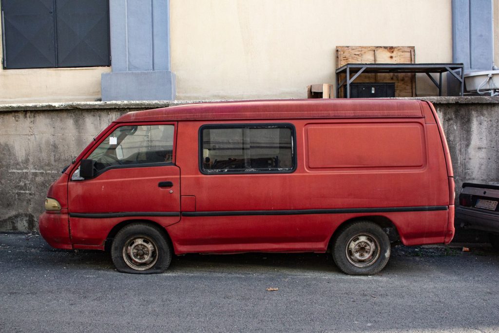Abandoned red van with flat tire parked on urban pavement.