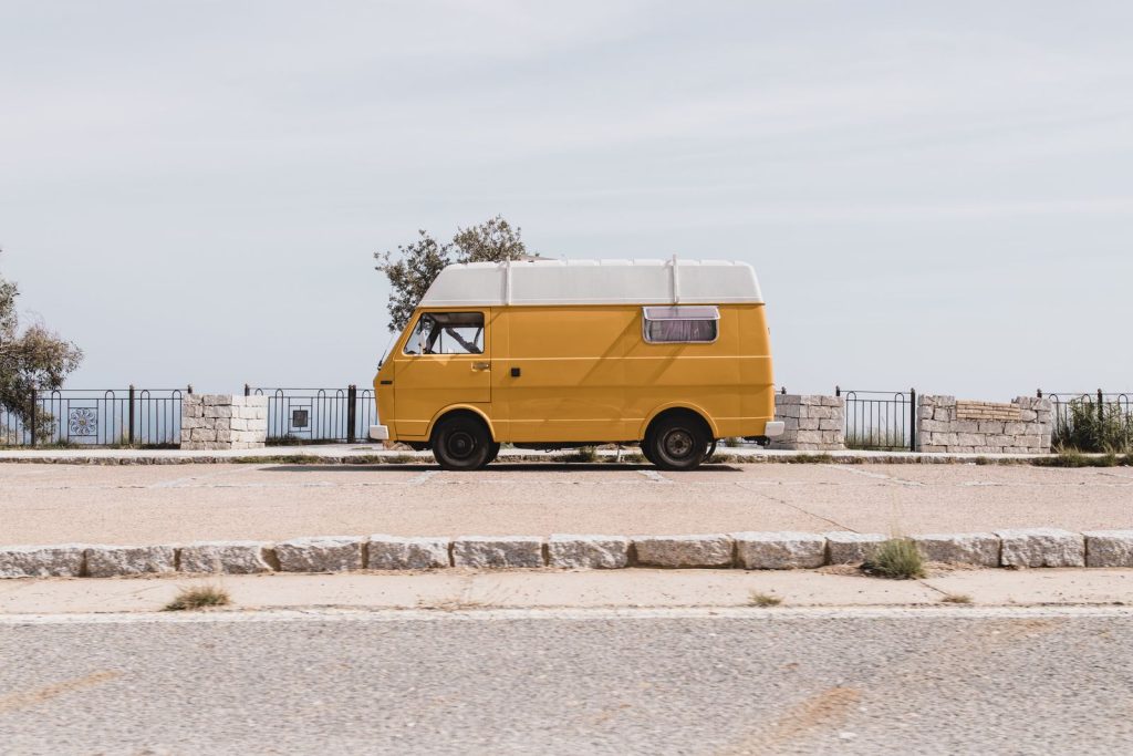 A classic yellow van parked by a scenic seaside road in Sardinia, Italy, under a clear sky.