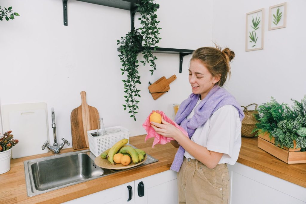 Side view of cheerful young woman in casual clothes standing and washing fruits in light kitchen in daytime