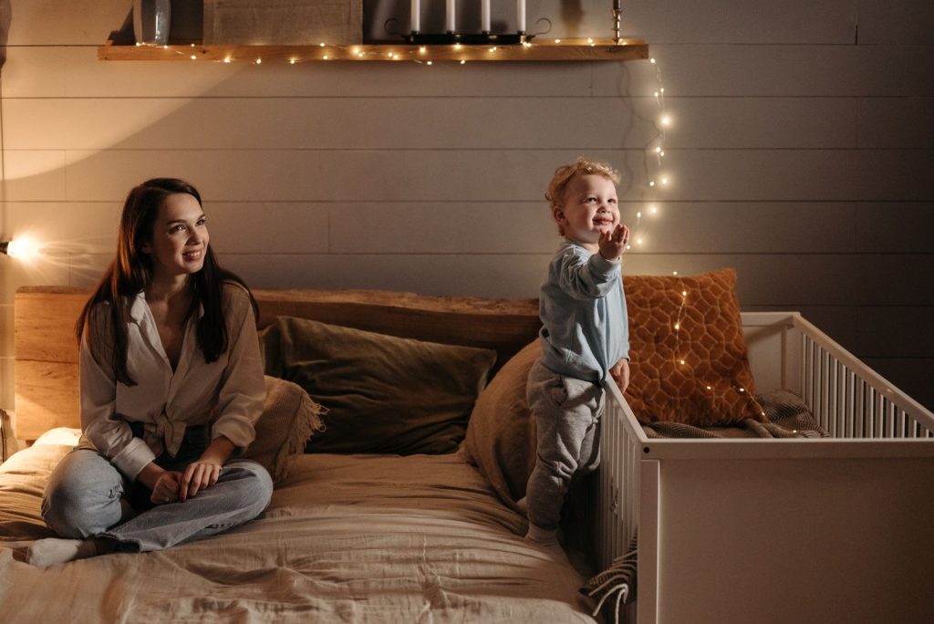 A mother smiles at her toddler son playing by the crib with warm lights.