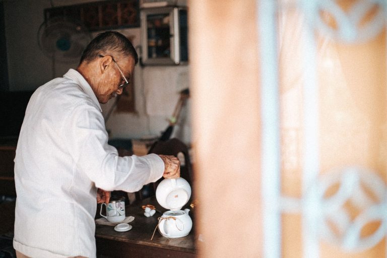 A senior man elegantly pours tea from a white teapot in a cozy room setting.